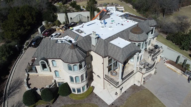 Wide drone view of white low-slope roof sections on a large stone home